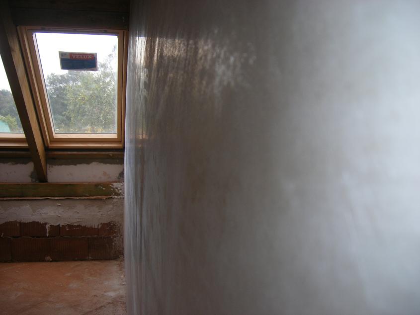 Świeżo nałożony, wilgotny tynk na ścianie poddasza, widoczne okno dachowe Velux i surowa cegła w tle.